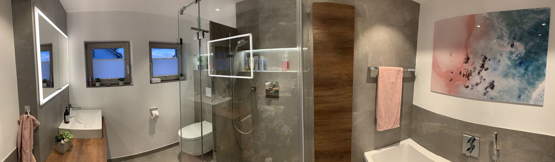 Panorama des neuen Bades: großformatige graue Fliesen, Holz-Waschtisch mit Aufsatzbecken, integrierter beleuchteter Spiegelschrank, bodengleiche Glasdusche mit beleuchteter Nische, wandhängendes Dusch-WC, in die Wand integrierter Holzschrank, Badewanne mit Unterputz-Armatur